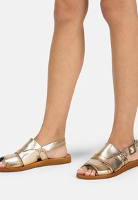 Sandales dorées métalliques à bouts ouverts, avec un design à bride réglable, semelle plate et texture lisse, dotées de brides minimalistes et d'une boucle.