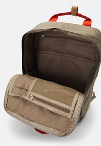 Tan backpack with red accents; features a main compartment, front mesh pocket, and inner zippered pocket for organization. Textured fabric.