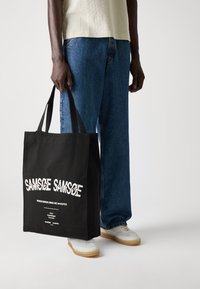 Black tote bag with white text, including "SAMSØE SAMSØE", held by a person in light top and blue jeans, featuring sturdy handles and a rectangular shape.