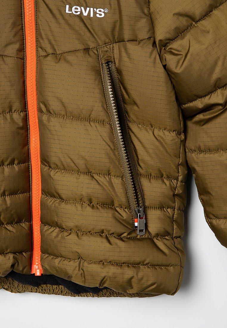 Veste matelassée vert olive avec texture matelassée, fermeture éclair orange et intérieur noir. Possède une poche latérale avec une fermeture éclair métallique.