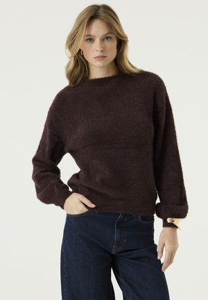 Vrouw met blond haar die een donkerbruine, pluizige trui en blauwe spijkerbroek draagt, staand met één hand in haar zak en de andere langs haar zij.