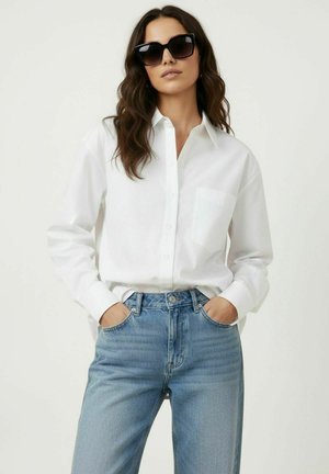 Femme portant des lunettes de soleil noires, une chemise blanche boutonnée et un jean bleu, debout, les mains dans les poches, devant un fond uni.