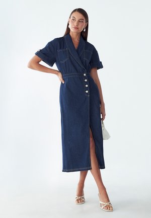 Frau steht mit der Hand in der Hüfte, trägt ein dunkles Denim-Midikleid mit kurzen Ärmeln, vorderen Knöpfen, Schlitz, weiße Sandalen und hält eine kleine weiße Tasche.