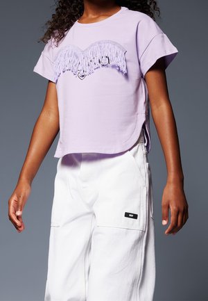 Pige iført en lavendelfarvet kortærmet top med hvidt hjerte med frynser og løse hvide bukser med sidelommer mod en grå baggrund.