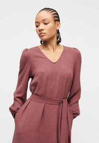 Γυναίκα με πλεγμένα μαλλιά που φοράει μακρυμάνικο μωβ φόρεμα με V-λαιμόκοψη και ζώνη στη μέση δεμένη, χέρια στις τσέπες, κοιτάζει αριστερά.