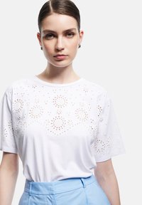 Hvid bomuldstrøje med dekorative laserskårne blomsterdesigns langs halsudskæringen og ærmerne, parret med lyseblå bukser.