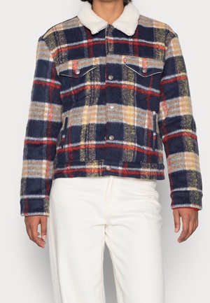 Person iført en navy, rød og beige ternet jakke med knapper, med en hvid sherpa-krave og hvide bukser, stående mod en ensfarvet baggrund.
