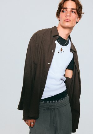 Brown checkered button-up shirt over a white sleeveless top, paired with grey pants. Silver necklaces and rings add detail.