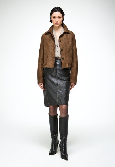 Femme portant une veste en daim marron, un chemisier beige, une jupe crayon en cuir noir, des collants transparents et des bottes noires pointues jusqu'aux genoux, debout devant un fond blanc.