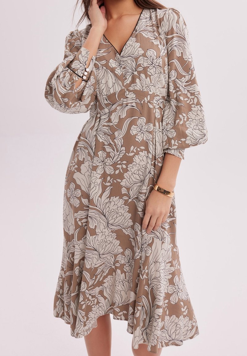 Femme portant une robe midi beige à imprimé floral blanc, manches bouffantes avec poignets boutonnés en perles, et un décolleté en V, posant sur un fond uni.