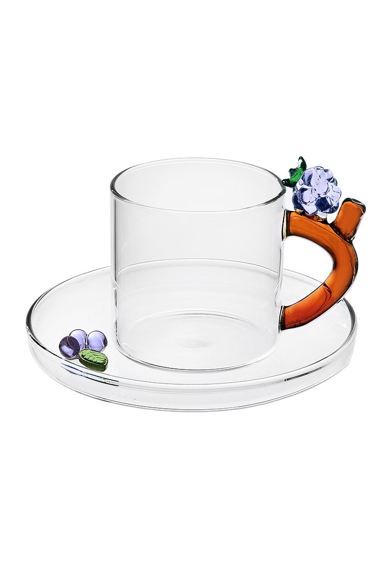 Przezroczysty szklany kubek z pomarańczowym uchwytem, ozdobiony fioletowym kwiatem i zielonymi liśćmi. Zawiera dopasowany, okrągły talerzyk z dekoracyjnymi koralikami.