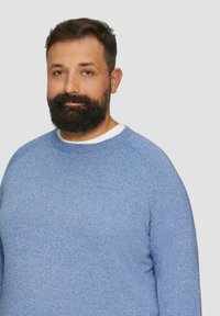 Hellblauer, gestrickter Pullover mit Rundhalsausschnitt, strukturiertem Muster und langen Ärmeln, getragen über einem weißen Hemd.