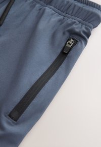 Grijze sportbroek gemaakt van zachte stof met een gladde textuur, voorzien van een zwart ritsvak aan de zijkant en een elastische tailleband.