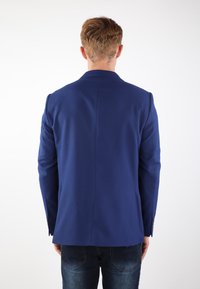 Blazer blu con un design strutturato, caratterizzato da un collo a punte, tessuto testurizzato e uno spacco posteriore singolo, abbinato a jeans scuri.