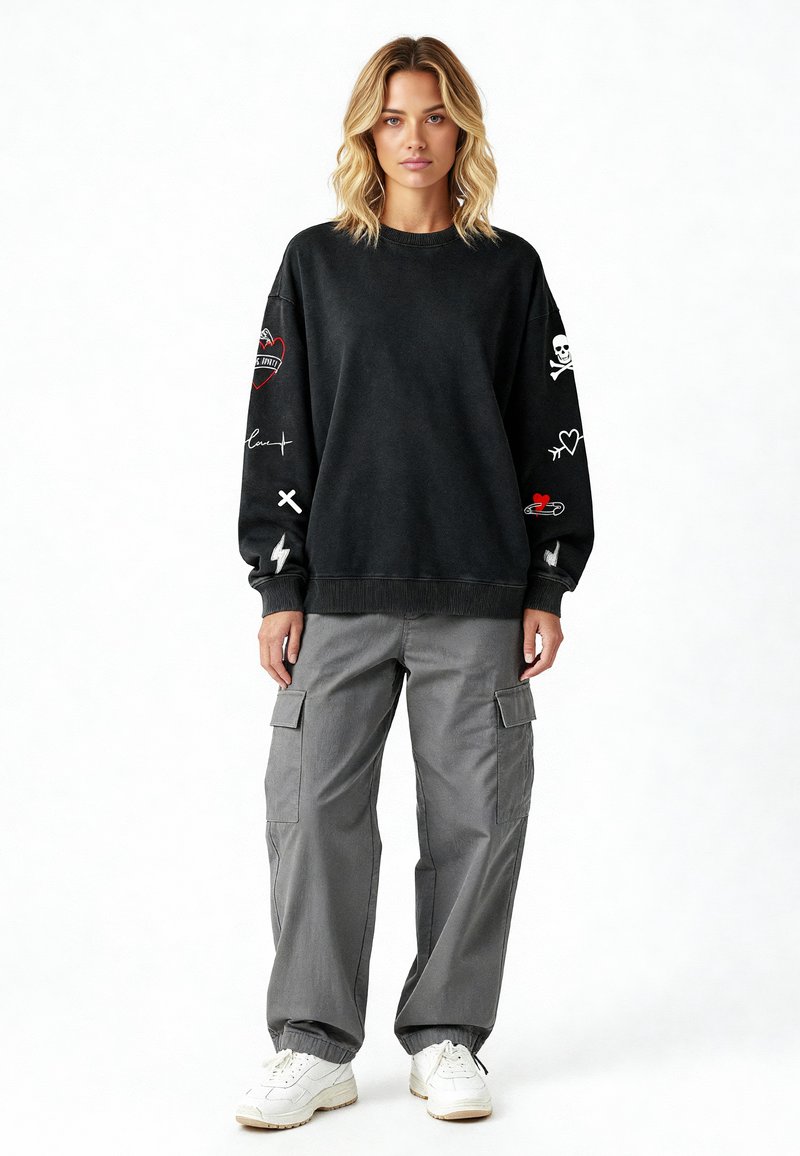 Sweat-shirt noir avec des graphismes brodés sur les manches, associé à un pantalon cargo gris avec des poches latérales et des poignets élastiques. Des sneakers blanches complètent le look.