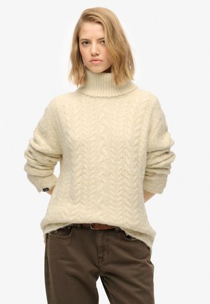 Cream cable-knit sweater with a high collar, loose fit, and rolled sleeves, worn over dark brown pants. Soft, textured fabric detail.