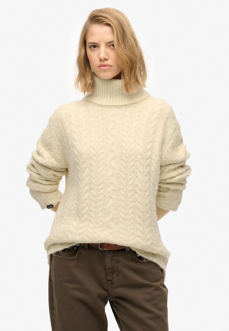 Cream cable-knit sweater with a high collar, loose fit, and rolled sleeves, worn over dark brown pants. Soft, textured fabric detail.