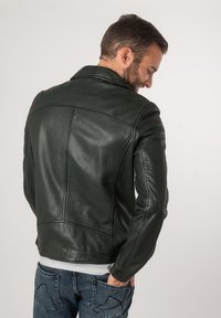 Giacca di pelle verde scuro con colletto strutturato, cuciture dettagliate e design aderente. Indossata sopra una camicia grigia, abbinata a un paio di jeans.