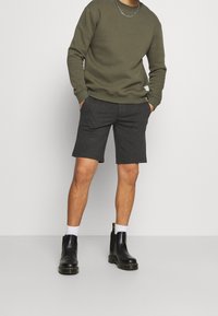 Felpa verde oliva con scollo rotondo, abbinata a pantaloni corti grigio scuro. Stivaletti neri alla caviglia con cuciture gialle e calzini bianchi in vista.