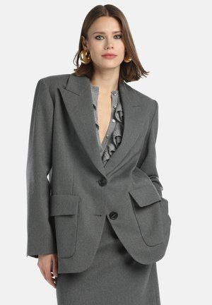 Woman wearing a gray oversized blazer with large pockets and a ruffled gray blouse underneath, paired with gold hoop earrings.