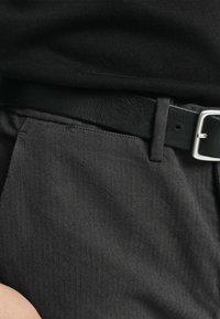 Zwarte leren riem met zilveren gesp, gedragen over donkergrijze stof in een visgraatpatroon. Close-up van het middelgebied.