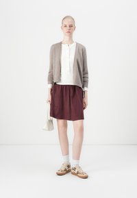 Lichtbruine cardigan over een witte top, bordeauxrode rok en gebloemde sneakers. Het model houdt een kleine crèmekleurige handtas vast, tegen een eenvoudige achtergrond.