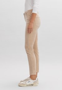 Pantaloni slim-fit beige chiaro con una trama liscia, design a cinque tasche e gamba leggermente svasata, abbinati a sneaker bianche con dettagli.
