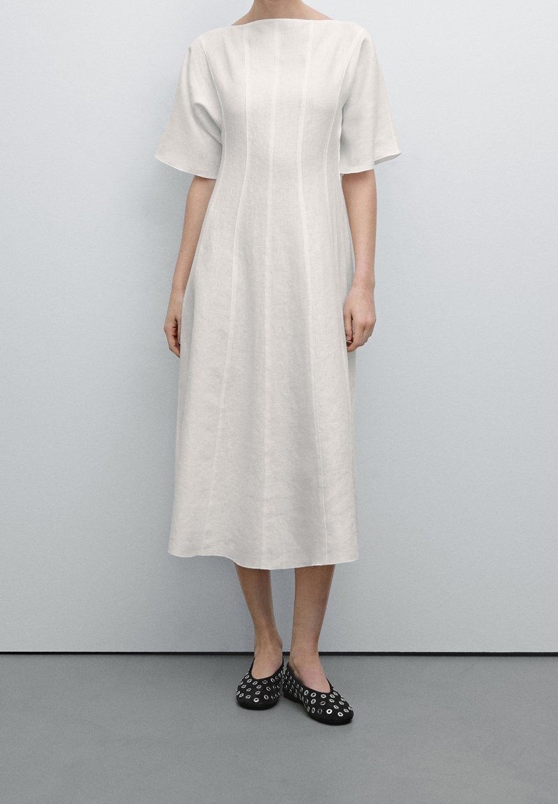 Femme portant une robe blanche jusqu'aux genoux à manches courtes et des chaussures noires à enfiler avec des motifs circulaires blancs, debout contre un mur clair.