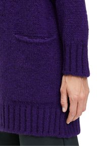 Lila stickad cardigan med ribbade manschetter och nederkant, med en ficka i patchwork. Den strukturerade tyget ser mjukt och tjockt ut.