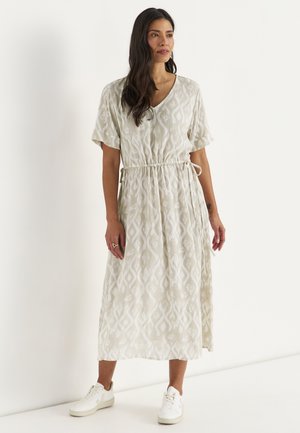 Frau mit langen dunklen Haaren, die ein hellbeiges gemustertes Midikleid mit kurzen Ärmeln und weißen Sneakers trägt, steht vor einem weißen Hintergrund.