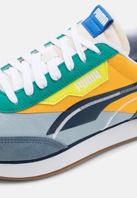 Farbiger Sneaker mit einer Mischung aus Türkis, Gelb und Grautönen, weißen Schnürsenkeln und einem blauen Logotag auf der Zunge.