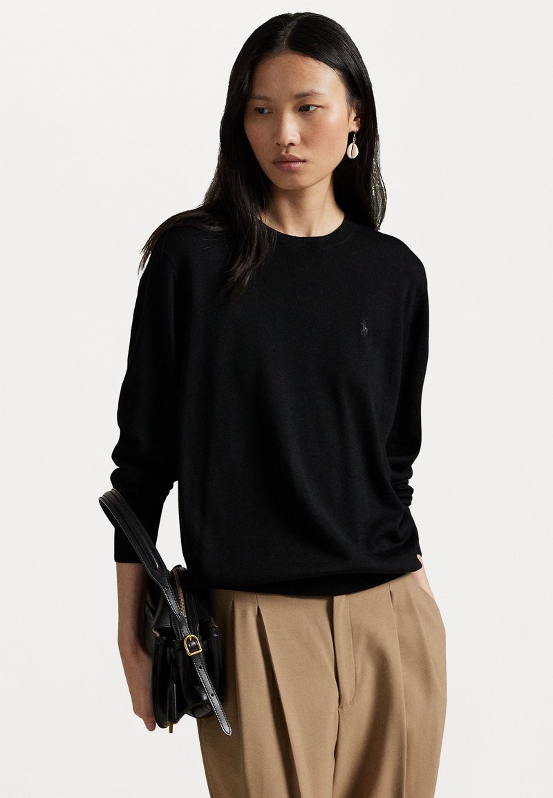 Black knit sweater with a round neck, paired with tan pleated trousers. Model holds a black leather bag with a gold-accented clasp.