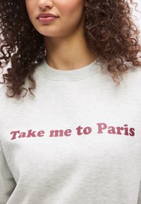 Γυναίκα με σγουρά μαλλιά που φοράει ανοιχτό γκρι φούτερ με το κείμενο «Take me to Paris» σε σκούρα ροζ γράμματα στο στήθος.