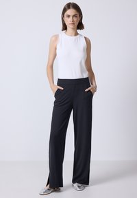Top bianco senza maniche abbinato a pantaloni larghi neri a coste con spacchi laterali. Scarpe argento slip-on completano il look.