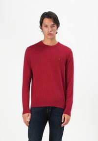 Pull en tricot rouge avec un col rond, des manches longues et un ourlet côtelé. Présente un petit logo sur la poitrine. Associé à un jean en denim foncé.