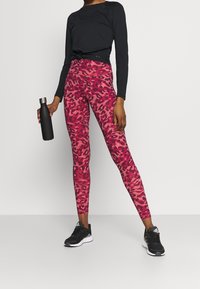 Person wearing black long-sleeve top and pink leopard-print leggings holding a black water bottle, standing against a plain white background.