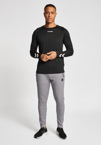 Cămașă sport neagră cu mânecă lungă și logo alb, completată de pantaloni joggeri gri, ajustați, cu un patch logo negru. Adidași negri.