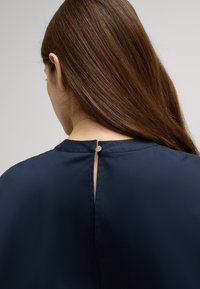 Marinebluse mit rundem Ausschnitt, Schlüssellochausschnitt hinten und einem kleinen silbernen Knopf. Glattes Material mit dezentem Glanz.