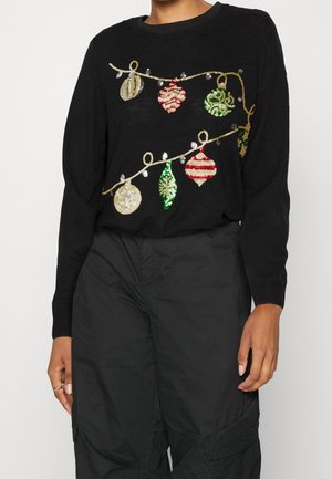 Personne portant un pull noir orné de décorations de Noël à sequins et de motifs en fil doré, associé à un pantalon noir. Seuls le torse et le haut des jambes sont visibles.