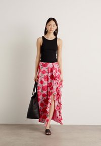 Débardeur noir, jupe maxi fleurie rose avec volants et fente, grand sac fourre-tout noir et sandales noires à glisser. Texture de tissu lisse.