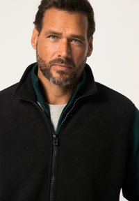 Schwarze Fleece-Weste mit einem Reißverschluss, hohem Kragen und einer inneren grünen Schicht, die eine glatte Textur aufweist und keine sichtbaren Verzierungen hat.