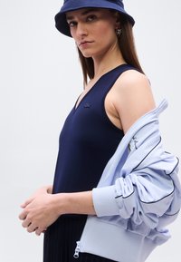 Jeune femme portant un haut sans manches bleu marine et un chapeau bob, tenant une veste zippée bleu clair avec des bordures noires sur un bras.