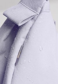 Vaaleanlila vedenpitävä laukku, jossa on teksturoitu kangas, pinnassa vesipisaroita, taitettu läppä ja näkyvä sauman yksityiskohta.