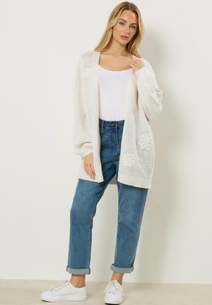 Mujer con cárdigan de punto blanco con estampado floral sobre camiseta blanca y vaqueros azules con puños, posando frente a un fondo liso y claro.