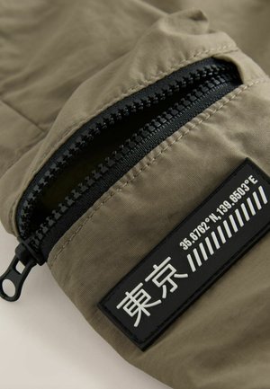 Olivenfarbener Stoff mit einer schwarzen Reißverschlusstasche, strukturiertem Akzent und einem rechteckigen Patch mit aufgedruckten Koordinaten und Symbolen.