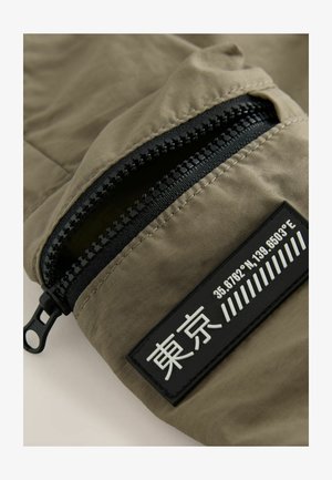 Kangaspala oliivinvihreää, jossa on mustalla vetoketjullinen tasku, teksturoitu yksityiskohta ja suorakulmainen läppä, jossa on painetut koordinaatit ja symbolit.