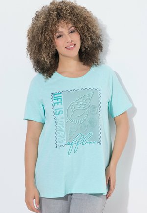 Lächelnde Frau in einem hellblauen T-Shirt mit Muschelmuster und dem Text „Das Leben ist offline schön“, stehend vor weißem Hintergrund.