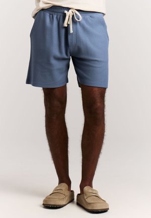 Man met lichtblauwe shorts met trekkoord en beige instaploafers, staand tegen een effen lichte achtergrond.