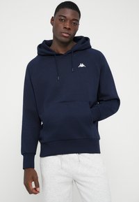 Νεαρός άνδρας φοράει μια ναυ blue μπλούζα με κουκούλα και μπροστινή τσέπη, μαζί με ανοιχτού γκρι φόρμες, μπροστά σε έναν απλό φόντο.