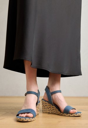 Pieds portant des sandales compensées en denim bleu avec des brides à la cheville, visibles sous une longue jupe noire, debout sur un sol en bois.